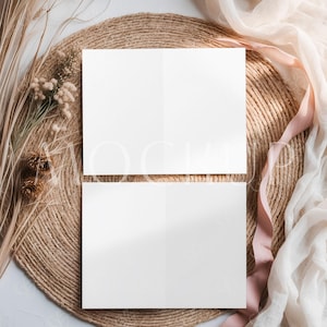 May include: Two blank white folded cards arranged on a woven, circular placemat. Dried grasses, small flowers, and pinecones are arranged to the left. A sheer, cream-colored fabric and a pink ribbon drape across the right side, creating a soft, neutral aesthetic.