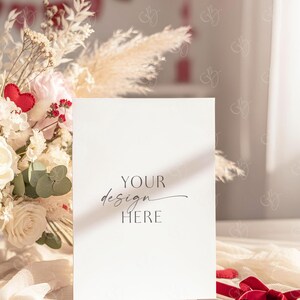 May include: A blank white card with the text "YOUR design HERE" is propped up in front of a floral arrangement. The arrangement includes white roses, pampas grass, and a red felt heart. A red ribbon and small felt hearts are in the foreground.