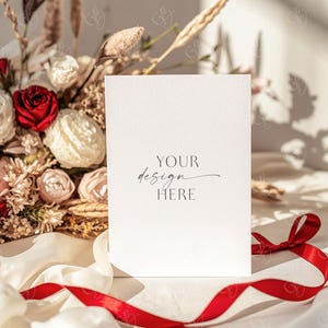 May include: A blank white card with the text "YOUR design HERE" in elegant script, resting on a white surface with a red ribbon. A bouquet of flowers with red and white roses and dried grasses is in the background.