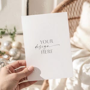 May include: A white card held by a hand, with the words "YOUR design HERE" in elegant script. The card is in front of a blurred background with a wicker chair and decorative elements, creating a soft, neutral aesthetic.