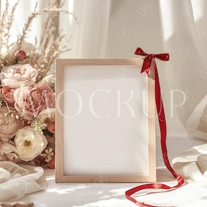 以下が含まれることがあります： 中央に「MOCKUP」の文字が入った木製のフォトフレーム。赤いリボンで飾られています。フレームは、柔らかい白い布とピンクとクリーム色の花のブーケを背景に配置されています。小さな赤いハートも存在します。