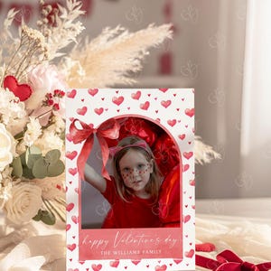 May include: A Valentine's Day card featuring a photo of a child in a red outfit, framed by a heart-patterned border. The card includes the text "happy Valentine's day" and "THE WILLIAMS FAMILY". A red ribbon and floral arrangement are in the background.