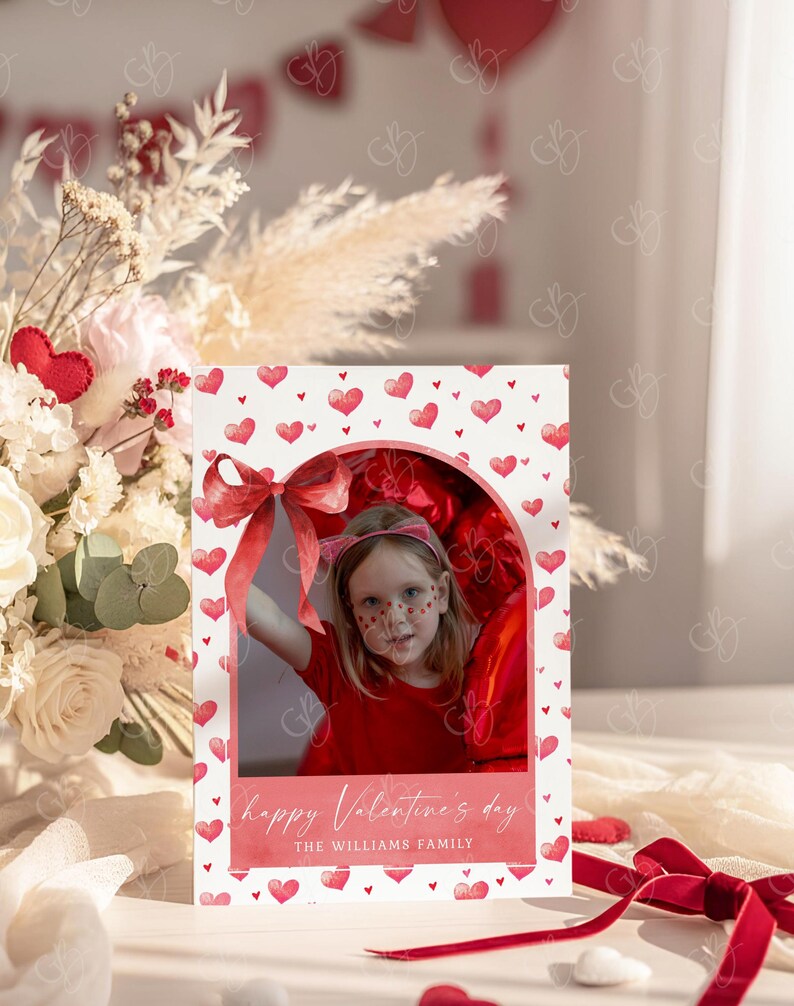 May include: A Valentine's Day card featuring a photo of a child in a red outfit, framed by a heart-patterned border. The card includes the text "happy Valentine's day" and "THE WILLIAMS FAMILY". A red ribbon and floral arrangement are in the background.