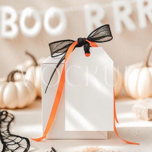 Könnte beinhalten: Eine weiße Geschenkbox mit einem orangefarbenen Band und einem schwarz-weißen Band, das darum gebunden ist. Ein weißes Etikett mit einer schwarzen Schnur ist an der Box befestigt. Das Etikett ist leer. Die Box steht auf einer weißen Oberfläche mit weißem Hintergrund. Im Hintergrund befindet sich ein Banner mit der Aufschrift "BOO" und "RRR".