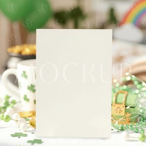 May include: A blank, off-white card stands on a white surface, surrounded by St. Patrick's Day decorations. A green treasure chest, shamrock-decorated mug, green beads, and a rainbow are visible. A green balloon and banner are in the background.