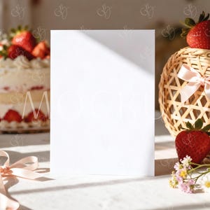 May include: A blank white card stands upright, illuminated by sunlight, with a strawberry cake and a woven basket with strawberries in the background. A pink ribbon and small flowers are also present, creating a soft, inviting scene.