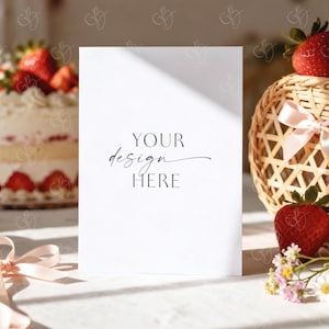 May include: A white card with the text "YOUR design HERE" is the focal point of this image. A strawberry shortcake, a woven basket with strawberries, and fresh strawberries are arranged around the card. Soft light and shadows create a delicate, inviting scene.