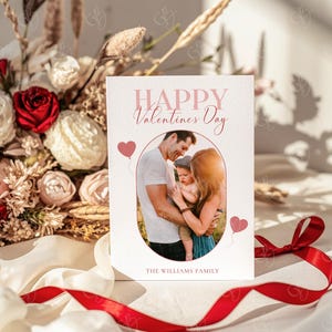 May include: A Valentine's Day card with a family photo inside an oval frame. The card reads "Happy Valentines Day" in pink script, with two pink heart balloons. A red ribbon and a bouquet of flowers are in the background.