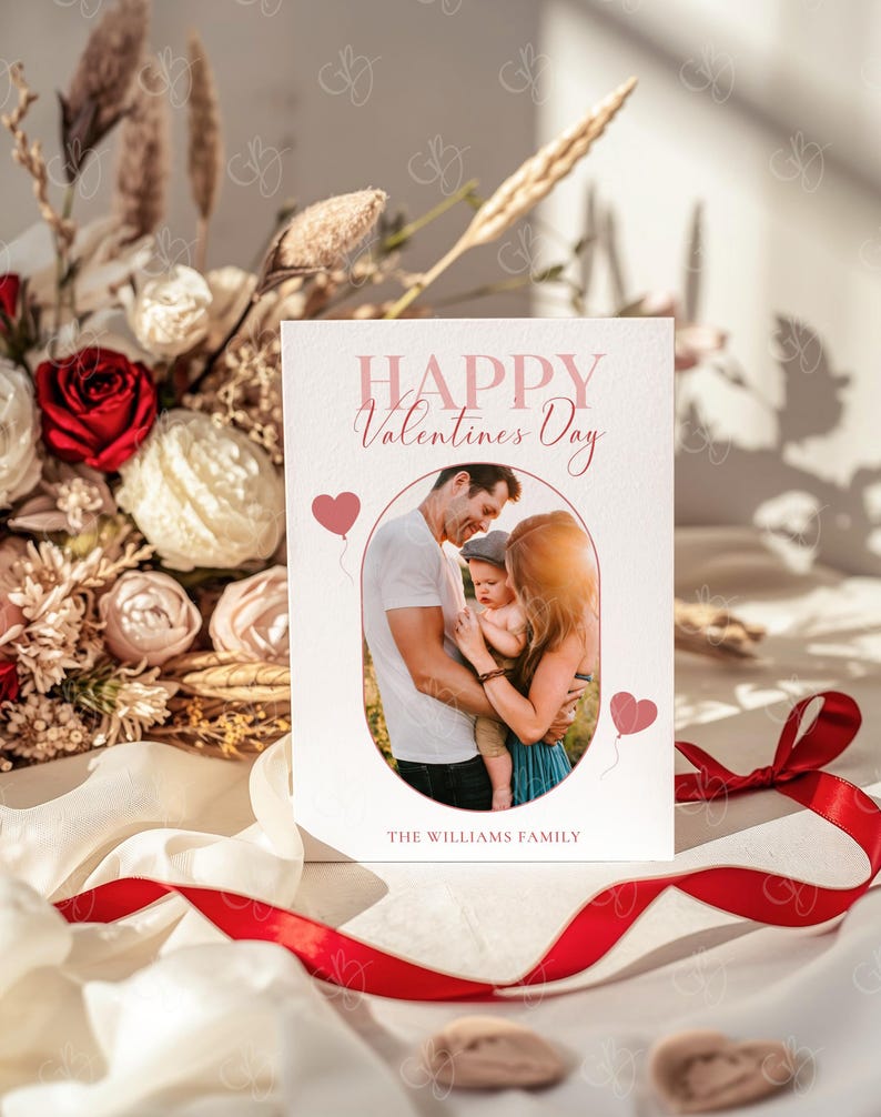 May include: A Valentine's Day card with a family photo inside an oval frame. The card reads "Happy Valentines Day" in pink script, with two pink heart balloons. A red ribbon and a bouquet of flowers are in the background.