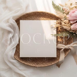 May include: A square, cream-coloured book with the word "MOCKUP" printed on the cover, placed on a woven, circular tray. A bouquet of pink flowers and dried grasses is to the right, with a cream-coloured ribbon.