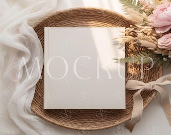 May include: A square, off-white book with the word "MOCKUP" printed on the cover, resting on a woven, circular tray. A bouquet of pink flowers and dried grasses sits to the right, with a cream-colored ribbon.