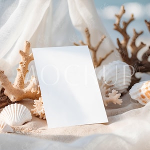 Puede incluir: Una tarjeta blanca en blanco se encuentra sobre arena, rodeada de conchas marinas, coral y una tela blanca transparente. El fondo presenta un océano y un cielo borrosos, creando una maqueta con temática de playa.
