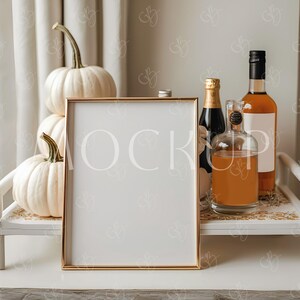 May include: A gold-framed blank sign stands on a white tray with bottles of liquor and a champagne bottle. Two white pumpkins are stacked on the left. The tray has a gold floral design. The background includes a white curtain.