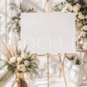 Pode incluir: Um cavalete de madeira com uma tela branca em branco, ideal para exibir obras de arte ou sinalização. Um vaso dourado com rosas brancas e vegetação está em frente ao cavalete. A palavra "MOCKUP" é visível na tela.