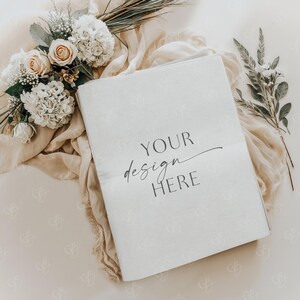 Puede incluir: Una tarjeta blanca con el texto "YOUR design HERE" sobre un fondo beige con un ramo de flores blancas y crema y follaje verde.