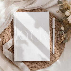 Puede incluir: Un libro blanco con la palabra «MOCKUP» impresa en la portada, colocado sobre un salvamanteles redondo tejido. Un bolígrafo blanco descansa junto al libro. La escena está decorada con una cinta blanca y flores secas.