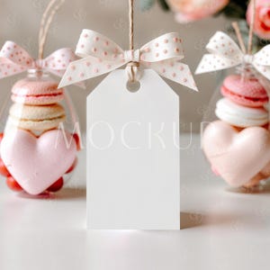 Könnte beinhalten: Ein weißes Geschenkanhänger mit einer rosa Schleife mit weißen Punkten, die an einem durchsichtigen Glasgefäß hängt, das mit rosa und weißen Makronen und einem rosa herzförmigen Bonbon gefüllt ist.