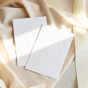 May include: Two blank white rectangular cards rest on a cream-colored fabric, with a cream-colored ribbon and dried wheat stalks. The cards are angled towards each other, creating a visually appealing composition. The scene is bathed in soft, natural light.