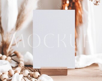 May include: A white rectangular card with the word "MOCKUP" faintly printed on it, resting on a small wooden block. The card is on a wooden table, with decorative beads and pampas grass in the background.