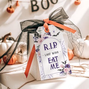 May include: A white gift tag with a purple floral design and a black spider web. The tag has the text "RIP - Last Wish - Eat Me" in purple lettering.