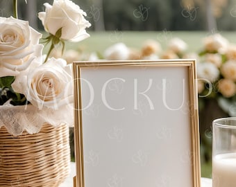 May include: A gold-framed rectangular sign with a white interior stands on a table. A wicker basket with white roses is to the left, and a glass with a white liquid is to the right. The background is blurred with greenery.