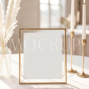 Peut inclure: Un cadre doré avec une feuille de papier blanche affichant le mot « MOCKUP ». Deux bougies blanches allumées dans des bougeoirs dorés se trouvent à droite. Un vase avec de l'herbe de la pampa séchée est à gauche.