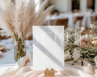 May include: A blank white card in a gold-colored holder, set on a table with a cream-colored satin runner. The table is decorated with pampas grass in a glass vase and a floral arrangement with pink roses.