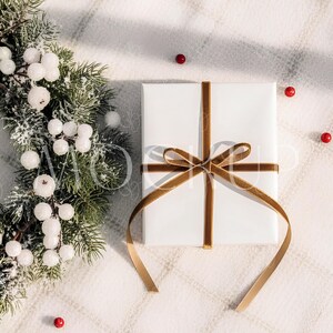 May include: A white gift box tied with a gold velvet ribbon, resting on a white, textured surface. A festive evergreen wreath with white berries and red berries are arranged nearby, creating a holiday theme.