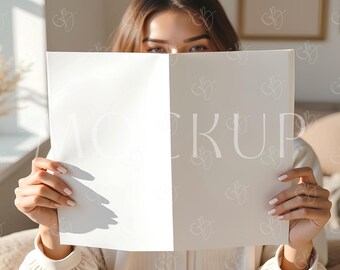 Puede incluir: Una persona sostiene un papel blanco con la palabra "MOCKUP" impresa. La persona lleva un vestido color crema. El fondo incluye un cuadro enmarcado y un interior de tonos neutros.