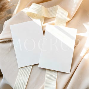 Op de afbeelding: Twee blanco witte rechthoekige kaarten met het woord "MOCK" erop gedrukt, rustend op een crèmekleurig lint. De kaarten en het lint zijn geplaatst op een gedrapeerde, crèmekleurige stof, met schaduwen van zonlicht.