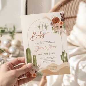 May include: A baby shower invitation with the words "Hello Baby" in a decorative script. The invitation features a desert theme with cacti, floral arrangements, and a neutral color palette. The details include the date, time, and location.
