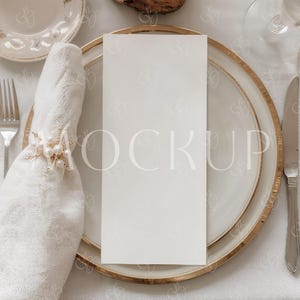 Op de afbeelding: Elegante tafelsetting met een witte menukaart op een goudomrande plaat. Een gevouwen witte servet wordt vastgehouden door een decoratieve gouden ring. Inclusief vork, mes en bloemaccenten.