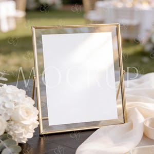 May include: A gold-framed sign holder with a blank white paper insert, ideal for displaying event information. The frame is propped up by a small stand. A white floral arrangement and cream-colored fabric are in the foreground, with a blurred outdoor setting in the background.