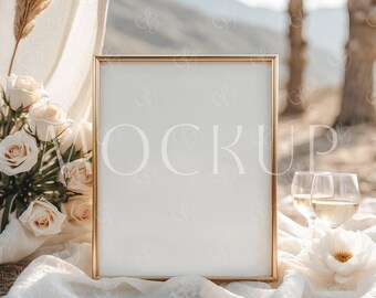 May include: A gold frame with a white background sits on a white fabric with white flowers and two glasses of wine. The frame is in front of a blurred background of palm trees and a desert landscape.
