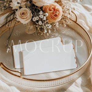 May include: A gold-rimmed tray holds white envelopes with gold edges, arranged near a bouquet of cream and peach-colored roses and other flowers. The word "MOCKUP" is faintly visible on the envelopes. The scene is set on a soft, neutral-toned fabric.