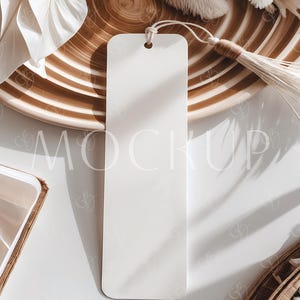 May include: A plain white bookmark with a string, set against a decorative wooden plate with brown and beige stripes. The background features white feathers and a white surface. The word "MOCKUP" is subtly visible.