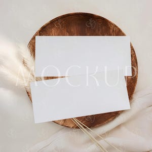 May include: A white mockup card with the word "MOCKUP" printed on it, resting on a wooden plate. The plate is set on a white cloth with a feather and some dried grass.