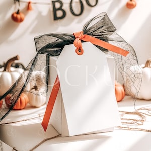 May include: A white gift box with a black and orange ribbon and a blank gift tag. The box is surrounded by small orange pumpkins and a black spiderweb. The background has a banner that says "BOO" in black letters.