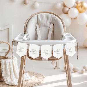 May include: A wooden high chair with a white fabric seat and a banner that says "YOUR design HERE" hanging from it. The banner is made of white cardstock with a brown twine string. The chair is decorated with a white and brown blanket and a wicker basket.