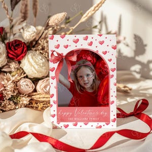 May include: A Valentine's Day card featuring a child in a red outfit with heart-shaped face paint. The card has a white background with red hearts and a red bow. Text reads "happy Valentine's day" and "THE WILLIAMS FAMILY."