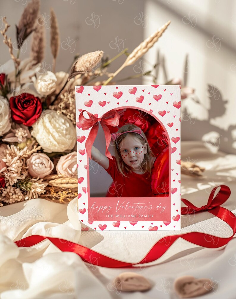May include: A Valentine's Day card featuring a child in a red outfit with heart-shaped face paint. The card has a white background with red hearts and a red bow. Text reads "happy Valentine's day" and "THE WILLIAMS FAMILY."
