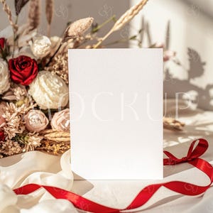 May include: A blank white card stands centered, with a bouquet of dried flowers and a red ribbon in the foreground. The bouquet includes red roses and cream-colored flowers. The ribbon is a vibrant red, and the background is softly lit.