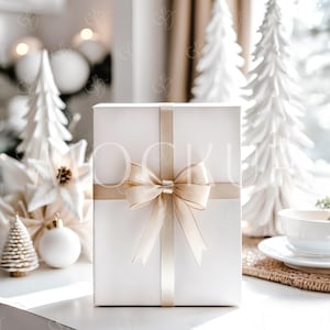 May include: A white gift box with a gold ribbon and bow is the focal point of this image. Decorative white Christmas trees, ornaments, and a poinsettia are in the background. A white teacup and saucer are on a woven placemat.