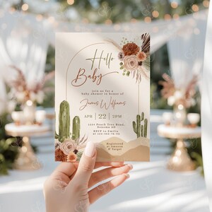 May include: A baby shower invitation with a desert theme. The invitation features a cactus, a floral arrangement, and the text "Hello Baby" in a handwritten font. The invitation also includes the date, time, and location of the shower, as well as the name of the guest of honor, James Williams. The invitation is registered at Amazon and Target.