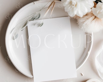 Può includere: Una scheda di carta bianca con la parola "MOCKUP" in testo grigio chiaro, posta su un piatto bianco. Il piatto è circondato da fiori bianchi, spighe di grano secche e un nastro bianco. Un rotolo di nastro adesivo si trova nell'angolo in basso a destra.