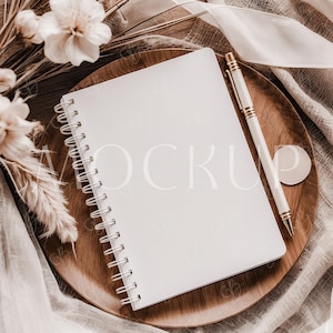 May include: A white spiral bound notebook with a blank page and a gold pen on a wooden plate with a white cloth background.