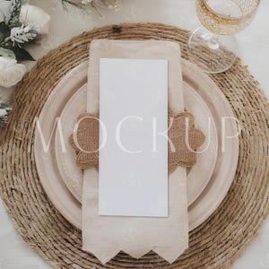 May include: A table setting with a woven placemat, a beige plate, and a white card. A beige napkin is placed on the plate, held by a burlap napkin ring. A decorative wine glass and floral arrangement complete the elegant, neutral-toned display.