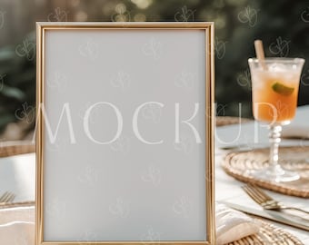 May include: A gold rectangular picture frame with the word "MOCKUP" in light gray text. The frame is on a table with a woven placemat, a glass of orange liquid with a straw, and a gold fork and knife.