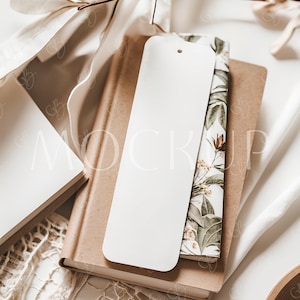 May include: A blank white bookmark rests on a stack of books with a floral patterned cover. The books are brown and beige, with a white background. A sprig of dried leaves and a white cloth add to the neutral, minimalist aesthetic.