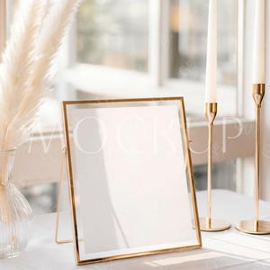 Peut inclure: Un cadre doré avec un intérieur blanc est posé sur une nappe blanche. Deux bougeoirs dorés avec des bougies blanches sont à droite. Un vase en verre transparent avec de l'herbe de la pampa séchée est à gauche. La scène est éclairée par la lumière naturelle.